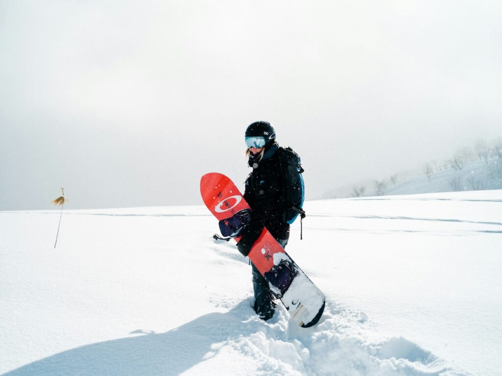 A snowboarder walking with her board in her hand while wearing her helmet, goggles, and neck gaiter.