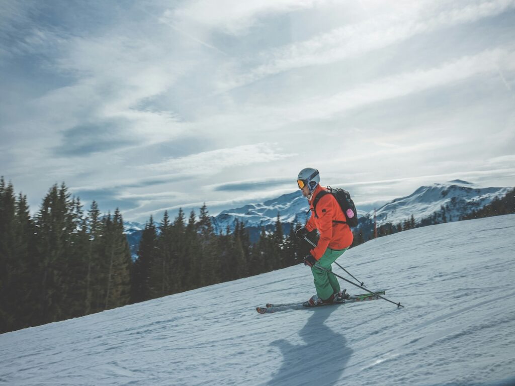 A man skiing down a slope with an orange jacket and green pants.