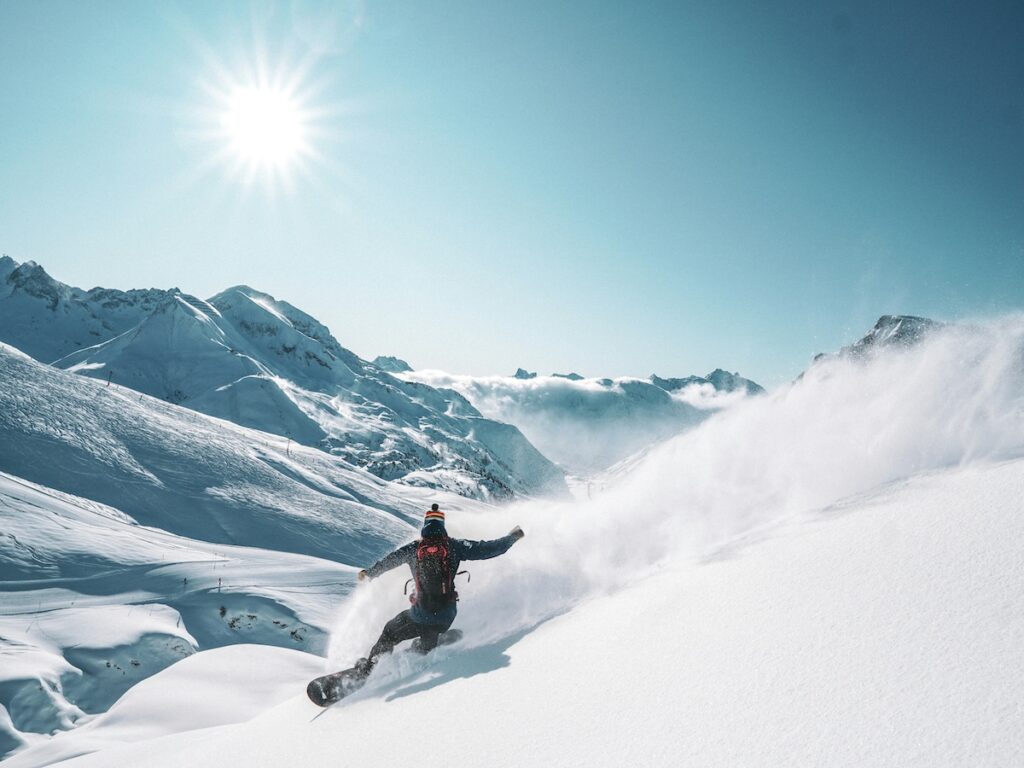 A snowboarder shredding down the slopes with mountains in the distance and sunny blue skies above.