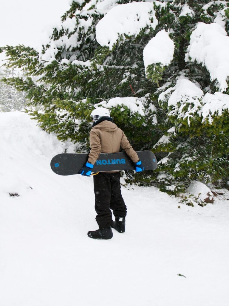 A snowboarder carrying his board beside a large tree.