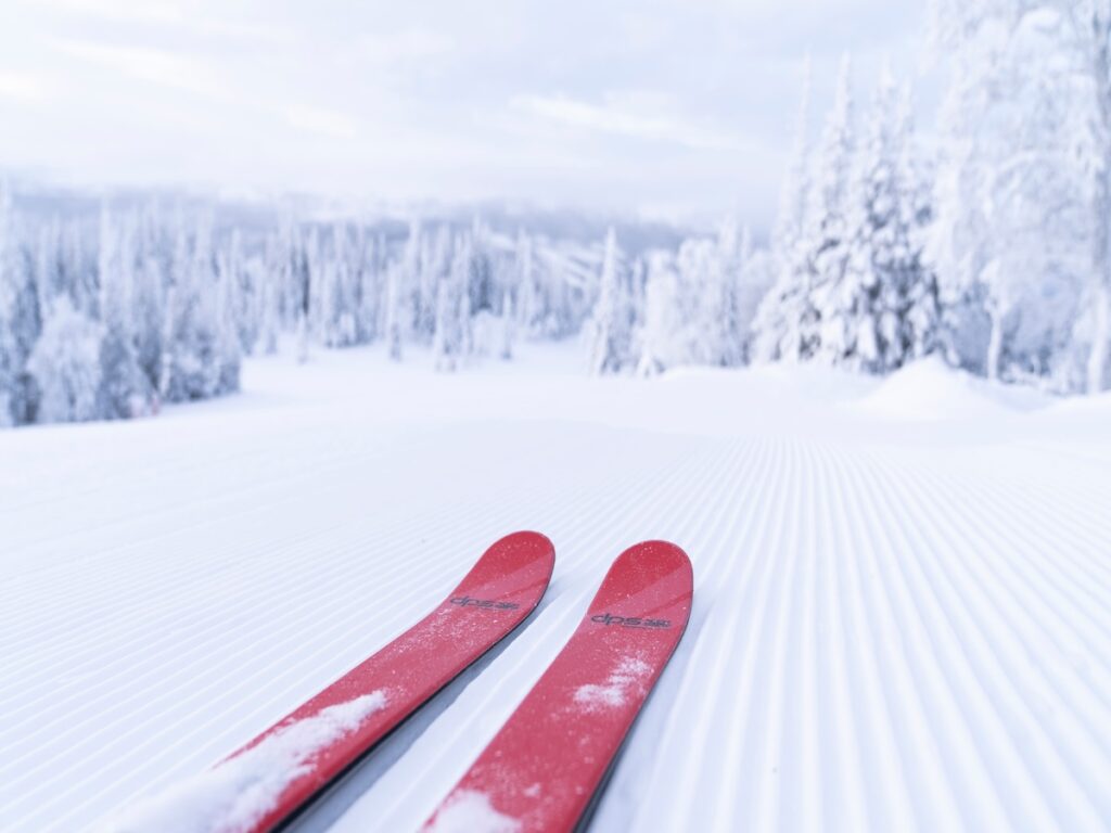 A pair of red skis on a groomed ski slope surrounded by snowy trees.