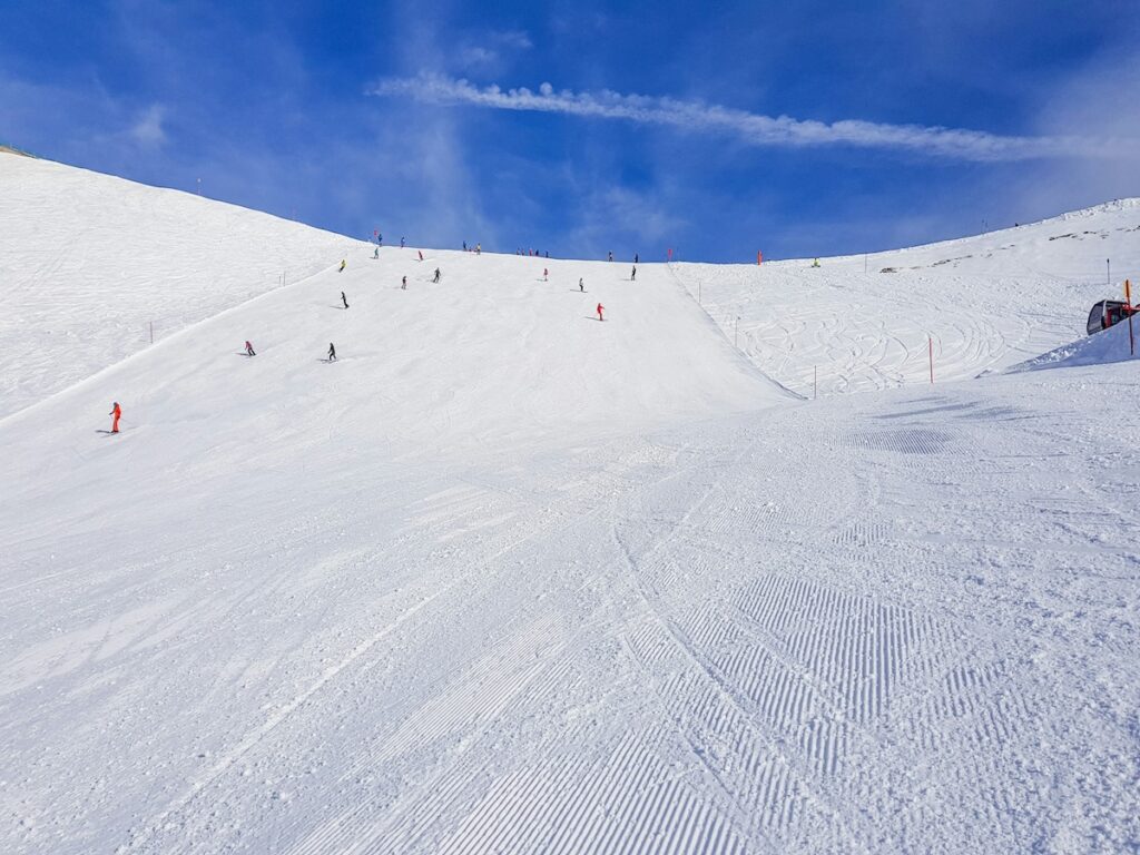 A bunch of skiers and snowboarders going down an easy slope on a bluebird day with a few small clouds.