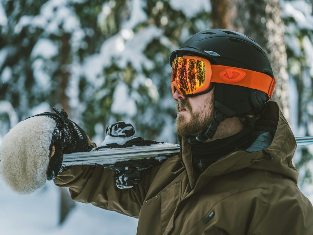 A male skier carrying his skis while he wear a pair of orange goggles.