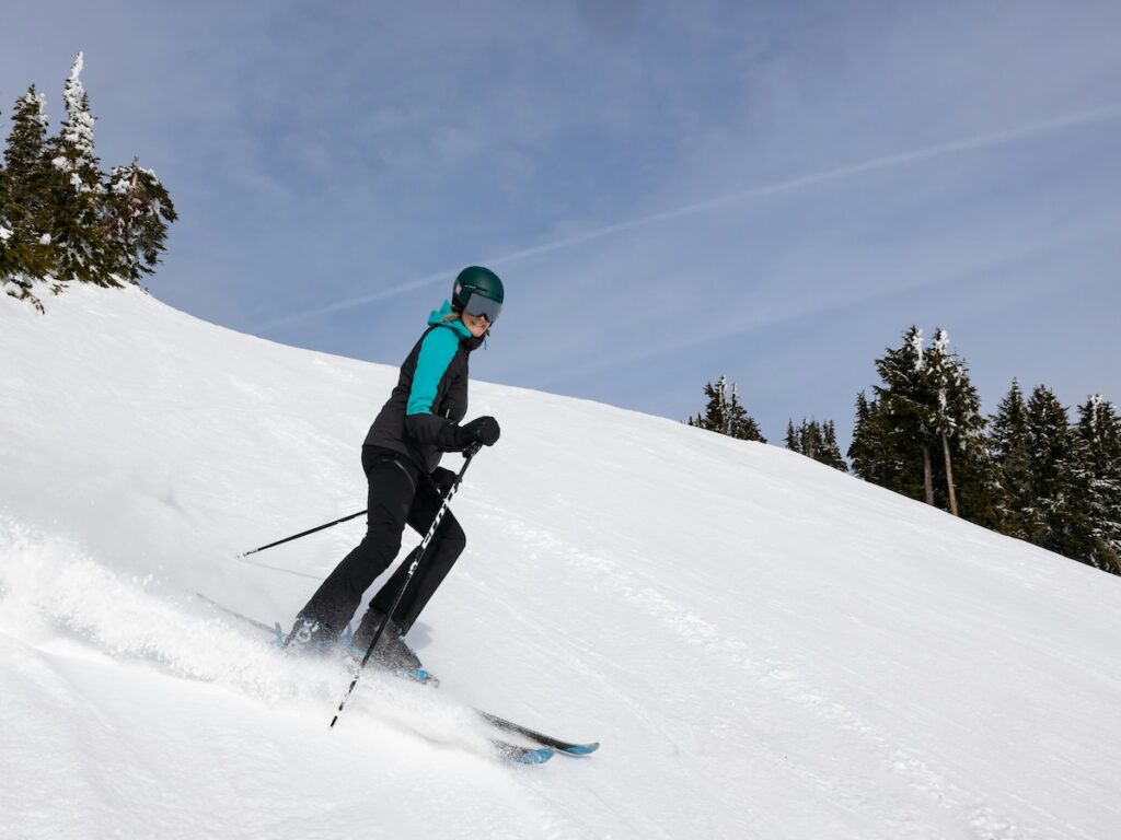 A skier smiling while going down the mountain wearing her hat, goggles, and gloves.
