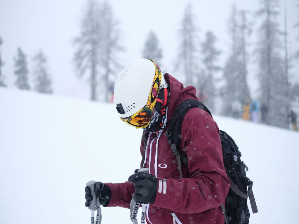 A skier in a maroon jacket looking down with their helmet and goggles on while it snows around them.