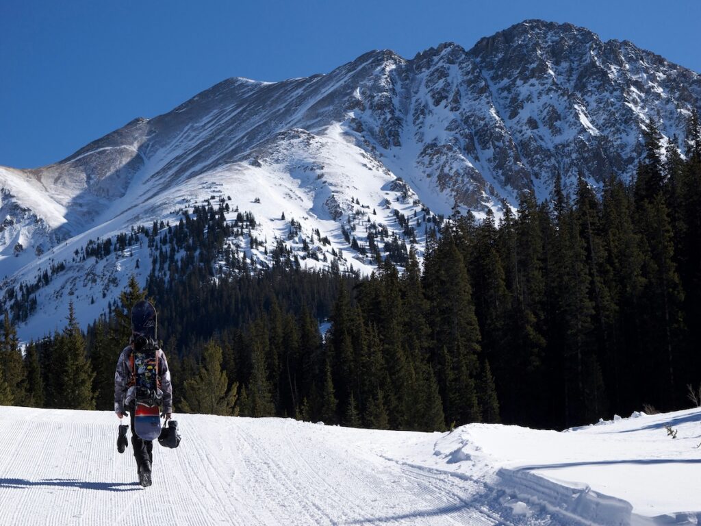A snowboarder carrying his board up a groomed run with mountains in the distance at Arapahoe Basin.