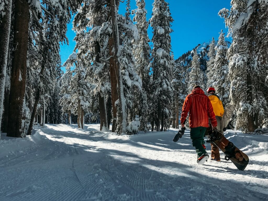 Two snowboarders carrying their boards at Mammoth Mountain through the trees.