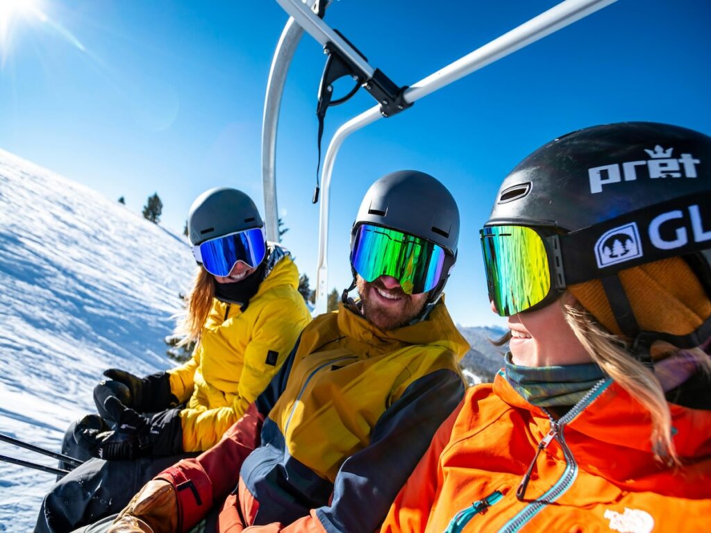 Three skiers smiling on the chairlift with goggles on and the sun shining in the distance.