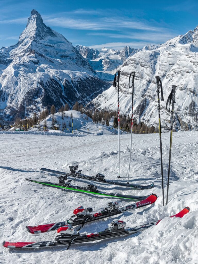 Two pairs of skis and two pairs of poles sitting in the snow with mountains behind them.