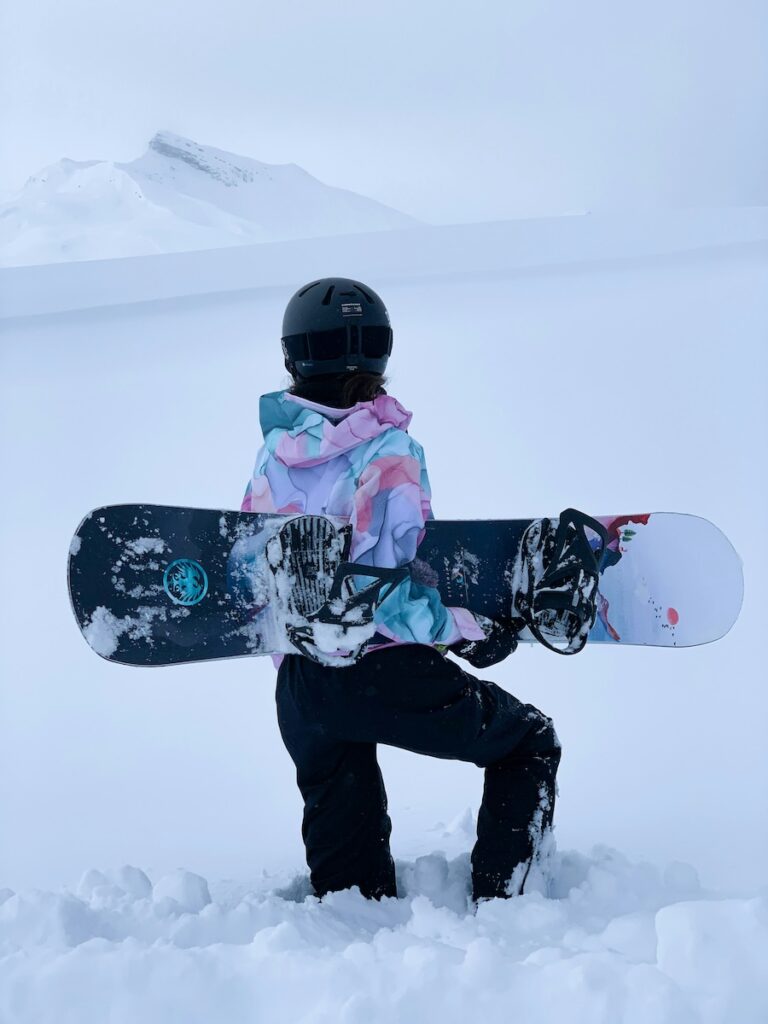A snowboarder wearing a colorful jacket, holding their board, and looking off in the distance.