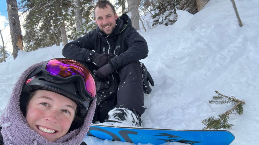 Abby and Sam smiling while sitting in the snow near some trees.