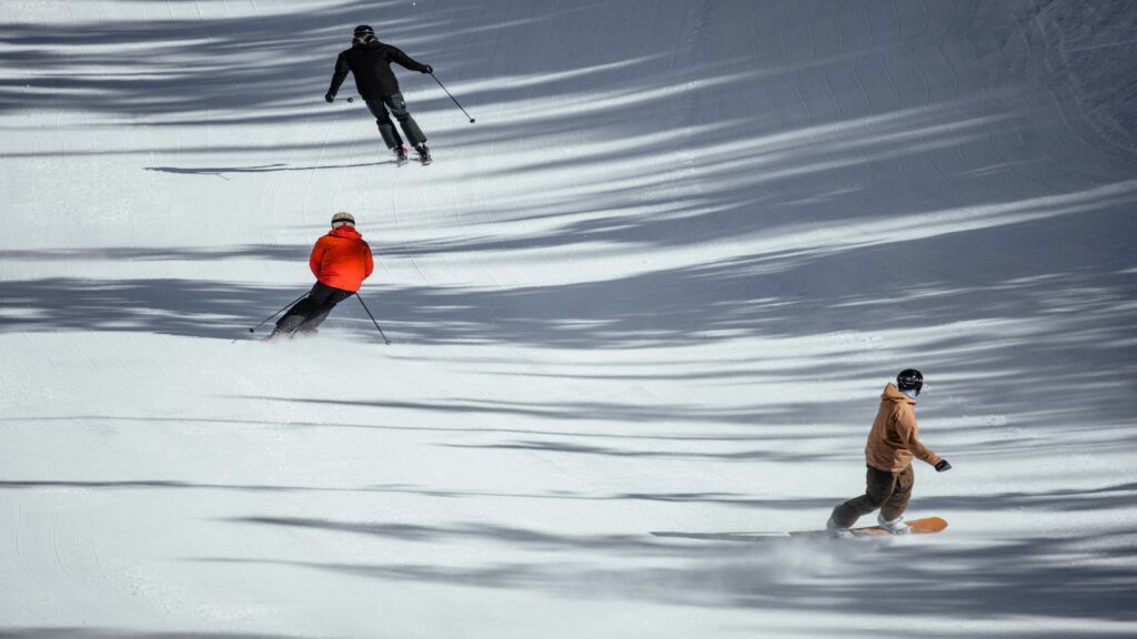 Two skiers and a snowboarder going down a groomed slope.