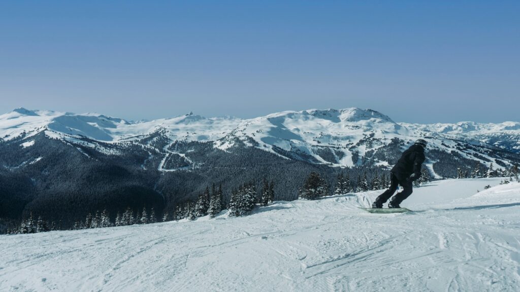 A snowboarder going down a groomed run at Whistler with mountains covered in trees off in the distance.