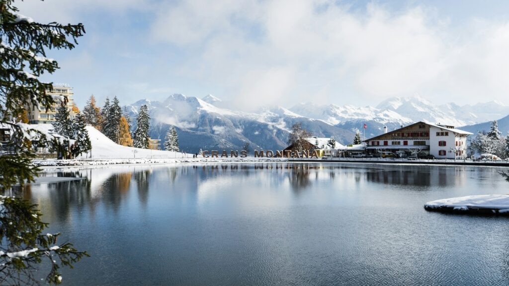 A Crans-Montana sign by a lake with snow covered mountains off in the distance. 